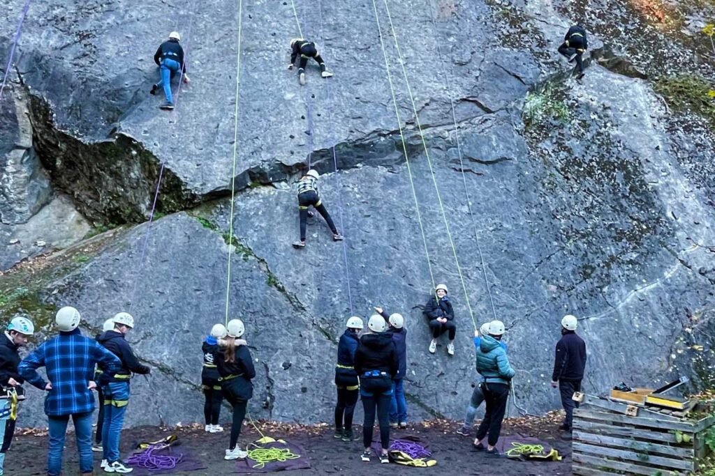 Een grote groep jongeren staat onder een klimwand of klimmen in harnas naar boven.