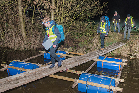 Kinderen lopen over een zelfgemaakte loopbrug met tonnen en sjorpalen over het water.