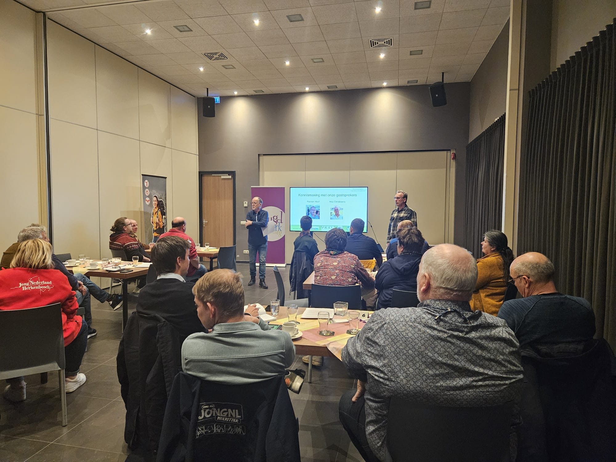 Een groep leiding luistert naar twee sprekers.