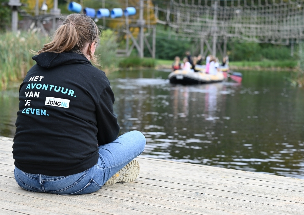 Een leidster zit op een vlonder en kijkt naar een groep leden op een rubberen boot op het water.